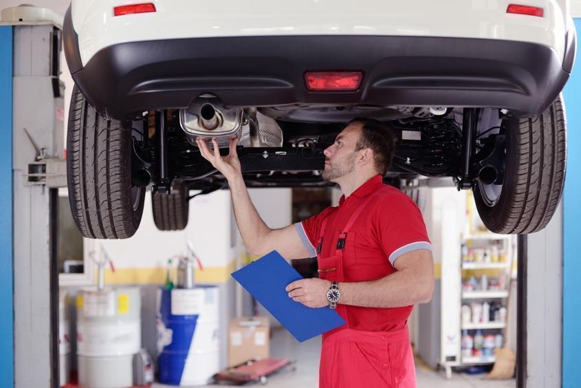 An automotive technician holding a clipboard while inspecting an exhaust system